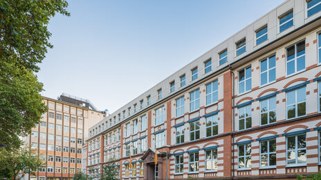 Foto des Altbaus und des Hochhauses an der Sonnenstraße mit Parkplatz davor. __ <br>View of the old building and the skyscraper on Sonnenstrasse across the parking lot.