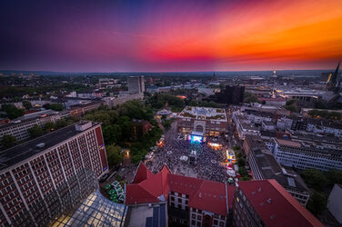 Die Luftbildaufnahme zeigt den Dortmunder Friedensplatz, auf dem zahlreiche Menschen vor einer Bühne stehen. Der Himmel hat einen Farbverlauf von bläulich bis gelblich.