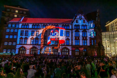 Auf eine Gebäude-Fassade ist eine Straßenbahn mit zwei Augen projiziert. Mehrere Menschen betrachten die Szene.