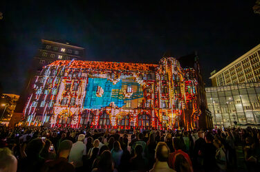 Auf eine Gebäude-Fassade sind durch eine Projektion die Buchstaben DO zu erkennen, mehrere Menschen verfolgen die Szene.