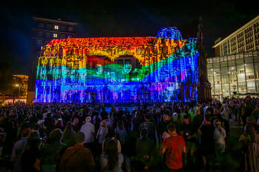 Eine Gebäude-Fassade ist in Regenbogenfarben angestrahlt, in der Mitte sind die Buchstaben DO zu erkennen. Mehrere Menschen verfolgen die Szene.