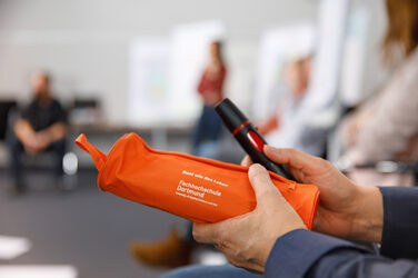 Nahaufnahme zweier Hände, die eine orangene Federmappe der Fachhochschule Dortmund und einen Textmarker halten.__Close-up of two hands holding an orange pencil case from Fachhochschule Dortmund and a highlighter.