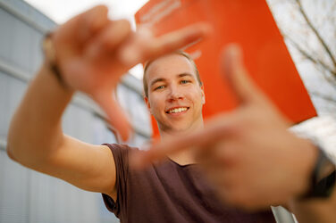 Foto eines Studenten, der mit Zeigefingern und Daumen vor sich ein Quadrat formt und dadurch guckt. .