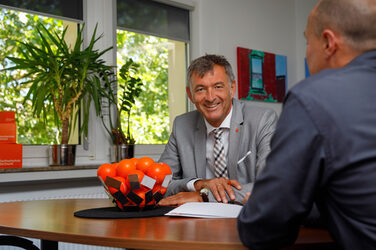 Foto des Kanzlers der Fachhochschule Dortmund, der am Tisch sitzt und in die Kamera lacht. Daneben ein weiterer Mitarbeiter in Rückansicht.__Photo of the chancellor of Dortmund University of Applied Sciences sitting at the table and laughing into the camera. Next to him, another employee in rear view.