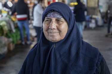 Eine Person trägt blaues Tuch am Oberkörper und über den Kopf gelegt. Ihr Gesicht ist freundlich, ihr Blick geht an der Kamera vorbei nach links. Hinter ihr ist eine Marktszene zu sehen.