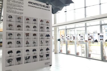 Das Bild zeigt ein erläuterndes Plakat im Foyer des Fachbereichs Architektur mit dem Titel "ARCHITECTS HELMET, STEGREIF BAUSTOFFTECHNOLOGIE" und bildet 36 verschiedenst gestalteten, bunten, plastisch und geometrisch stark veränderten Bauhelme ab. Im Hintergrund hängen erläuternde Plakate mit den dazugehörigen Helmen. Auf den Plakaten der Studierenden werden die wesentlichen Aspekte des Baustils eines gegebenen Architekten sowie die Materialwahl erläutert. Es wird außerdem ein Beispielgebäude vorgestellt, das zur Inspiration bei der Gestaltung des Helmes herangezogen wurde.