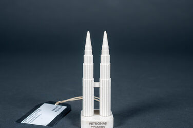 This picture shows a small model of two towers made of white plastic the size of a chess piece. The two towers are the same height, connected by a bridge about halfway up, and they stand on two square marble bases with strongly rounded corners and the name of the building as an inscription. The towers are the Petronas Towers in Malaysia, Indonesia. They look like two pens with vertical grooves, some levels of which are staggered from the tops downwards.