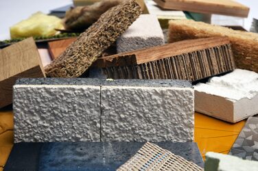 The picture shows a close-up of a disordered collection of different materials in a rectangular shape of the same dimensions with different orientations.
In the foreground of this picture are a solid, brown block of wood, a gray-black piece of polystyrene with a white plastered front, a composite piece made of a red-brown wooden panel with grooves, with a thick piece of cardboard glued to the underside, a fibrous, solid piece of brown wood fiber, a white, solid, porous piece of sand-lime brick, a thin, beige-colored mesh with two individual blue stripes in it, a white, solid piece of plaster that is very uneven on the surface, a fibrous, solid piece of brown wood fiber, a solid, smooth, shiny, dark blue-black marbled piece of rock and a solid, thin, patterned, silver-colored metal plate.
Behind it are other similar pieces of material of this type.