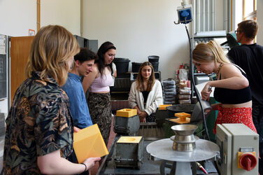 The picture shows a group of young people in the concrete laboratory of the Faculty of Architecture at the Fachhochschule Dortmund. On the table you can see the steel formwork molds in which some of the students have already placed their own cut Styrodur formwork for their self-designed sculptures. The orange-colored Styrodur formwork is held in the larger, cubic steel forms by clamping them together from the outside with screws. On the right-hand edge of the picture, a student is filling sand into the space between the steel formwork and her Styrodur formwork with a metal shovel to prevent the glued Styrodur formwork from being pushed apart when the concrete is poured in and the concrete from flowing out through gaps. Other students stand around and watch her work.