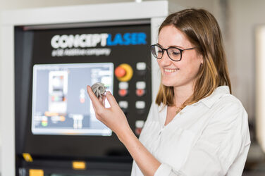 Photo of a woman looking at a 3D printed metal model in her hand, the printer can be seen out of focus in the background.