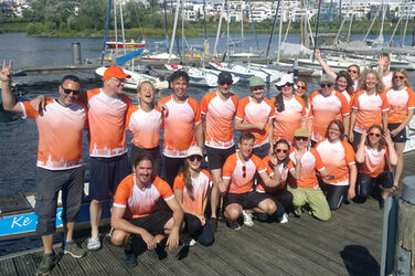 20 people in identical jerseys stand or squat on a jetty in front of a long canoe floating on the lake behind them.