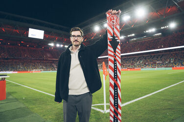 A person stands on the edge of the pitch in a stadium and presents a fan scarf.
