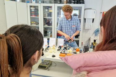 One person presents various objects on a table, two people watch.