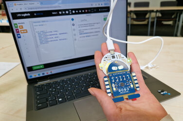 A hand holds a circuit board connected by cable in front of a laptop in the shape of a robot with illuminated LEDs.