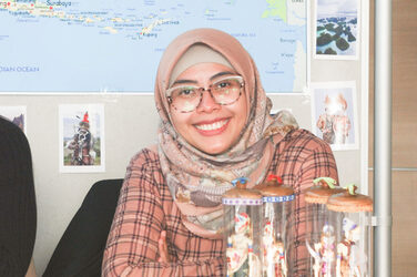 One of the exhibitors at the International Week's "Marketplace of Opportunities" sits smiling at a table with flyers. In the background is a map__One of the exhibitors at the International Week's "Marketplace of Opportunities" sits smiling at a table with flyers. In the background is a map