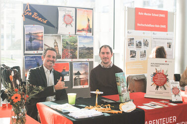 Ein weiterer Ausstellertisch des "Marktplatzes der Möglichkeiten": zwei Männer sitzen am Messestand und lächeln in die Kamera. Auf dem Tisch stehen orangene Blumen und Infomaterial__Another exhibitor table of the "Marketplace of Opportunities": two men sit at the booth and smile into the camera. On the table are orange flowers and information material