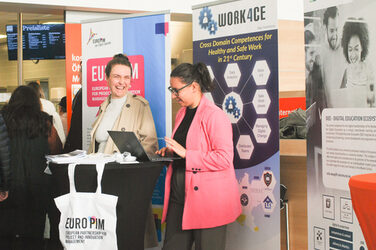 Zwei junge Frauen unterhalten sich lachend an einem Laptop an einem Messestand, beim "Marktplatz der Möglichkeiten"__Two young women are laughing and talking at a laptop at a booth, at the "Marketplace of Opportunities"