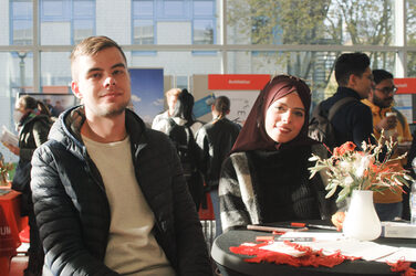 Eine junge muslimische Frau und ein junger Mann lächeln im Sonnenlicht an einem Tisch eines Messestands in die Kamera, beim "Marktplatz der Möglichkeiten"__ A young Muslim woman and a young man smile into the camera in the sunlight at a table of a booth, at the "Marketplace of Opportunities"