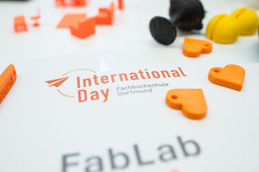 Poster des international Day umgeben von orangfarbenen Gegenständen aus dem 3D Druckers. International Day poster surrounded by orange-colored objects produced by the 3D printer.