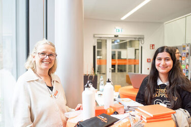 Eine Kollegin des International Offuce und eine Studierende stehen an einem Tisch und lächeln sich über die Gewinne eines Gewinnsspiels, dass der international Day veranstaltet. An employee and a student stand at a table and enjoy the prizes from a competition organized by International Day.