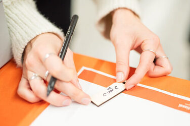 Auf einem orangfarbenen Tisch schreibt eine Person ihren Namen auf Georgisch auf einen Schlüsselanhänger aus Holz.On an orange-coloured table, a person writes their name in Georgian on a wooden key ring.