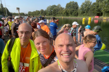 Selfie of three people under an open summer sky with many other people in the background on the bank of a watercourse.