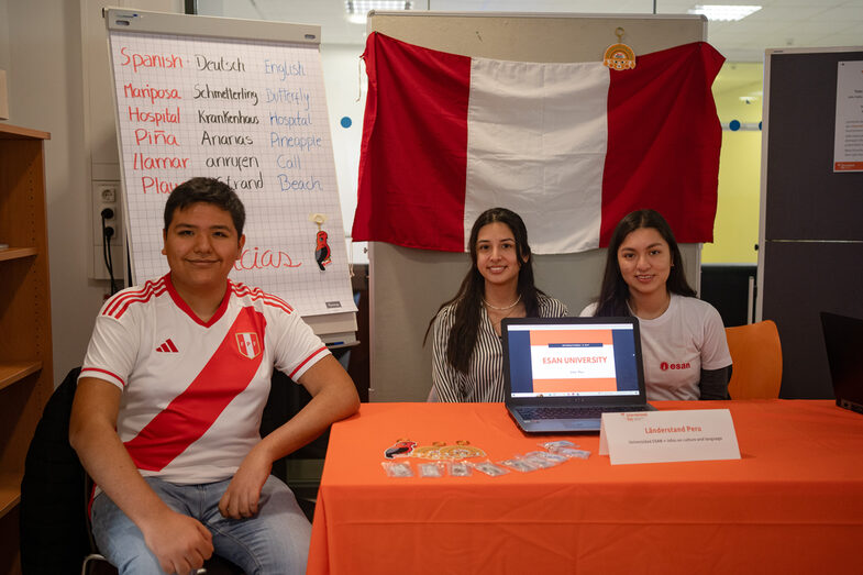 Three people from the Peru country stand are sitting at their stand and smiling at the camera __Three people from the Peru country stand are sitting at their stand and smiling at the camera.