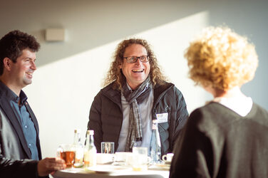 Drei Personen sind im Gespräch an einem Stehtisch im Sonnenschein, beim Welcome Event der Internationalen Woche__Three people are talking at a table in the sunshine, at the welcome evnent of the interational week