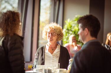 Drei Personen sind im Gespräch an einem Stehtisch im Sonnenschein, beim Welcome Event der Internationalen Woche.Im Hintergrund sind Fenster__Three people are talking at a table in the sunshine, at the welcome evnent of the interational week.In the background are windows