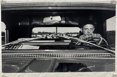 Schwarz-Weiß-Foto einer Person am Lenkrad eines Autos, durch die Windschutzscheibe gesehen.