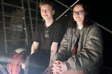 Two people sit on a kind of scaffolding platform with ladders in the dark background and look into the camera in a friendly manner.