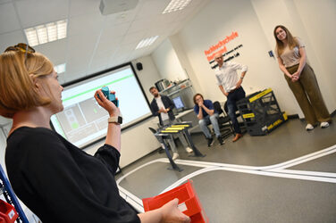 A person is standing in a laboratory holding items. She is explaining something. Other people are listening to her.