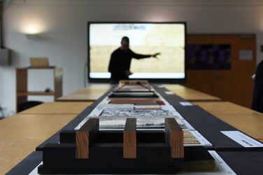 In the foreground, different materials from the exhibition can be seen, while in the background, blurred, the guest speaker gives his lecture in front of a monitor