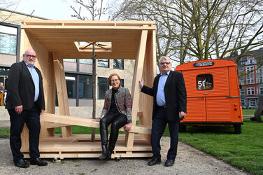 Eine weiblich gelesene Person sitz in einer Würfel-Holzkonstruktion. An den Ecken stehen zwei männlich gelesene Personen. Rechts daneben steht ein orangefarbener Citroen HY.