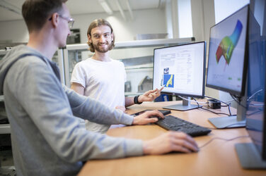 Two students are standing at a desk in front of two monitors showing FEM simulations of a motorcycle fork.