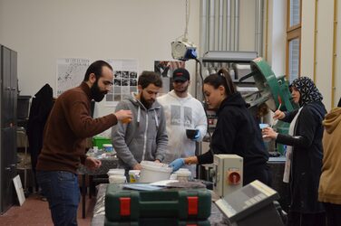 Sechs Studierende arbeiten an einem Werktisch im Betonlabor. Gemeinsam mischen sie mit Utensilien wie Mischbehälter und Rührstab Sprachtemasse an.