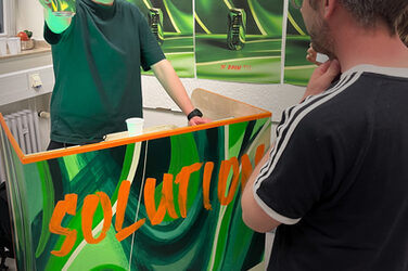 Eine Person hinter einem Stand mit der Aufschrift "Solution" hält eine Getränkedose in die Kamera.
