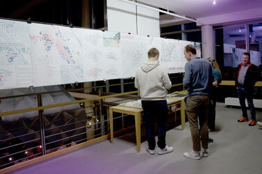 Mehrere Personen betrachten im Obergeschoss einer Ausstellung großformatige Pläne und Modelle zu städtebaulichen Projekten. Der Raum ist modern beleuchtet und wirkt wie Teil einer Präsentation oder Feier.