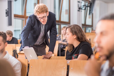 Foto eines Dozenten, der im Hörsaalgang steht und einer Studentin etwas auf ihrem Laptop zeigt. Drumherum unscharf weitere Studierende.__Photo of a lecturer standing in the lecture hall corridor showing a student something on her laptop. Around it blurred other students.