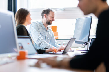 Foto von einer Mitarbeiterin und einem Mitarbeiter der Hoschschul-IT, die gemeinsam auf einen Computer-Bildschirm schauen. Im Vordergrund unscharf im Anschnitt ein weiterer Mitarbeiter am PC.__Photo of a female employee and a male employee of the IT Service of the Fachhochschule Dortmund, looking together at a computer screen. In the foreground, out of focus, another employee at the PC. <br><br>