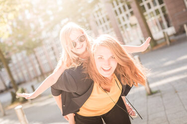 Foto einer Frau mit einem kleinen Mädchen, das sie huckepack trägt. Das Mädchen trägt eine orangene Sonnenbrille. __ A woman piggybacks a little girl. The girl is wearing orange sunglasses.