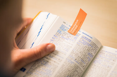 Foto eines aufgeschlagenen Wörterbuchs, das jemand in der Hand hält. Die geöffnete Seite ist mit einem orangenen Zettel mit FH-Wortmarke markiert.__Photo of an open dictionary held in someone's hand. The open page is marked with an orange slip of paper with the word mark of Fachhochschule Dortmund.