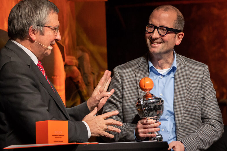 Foto von zwei männlichen Personen, die miteinander sprechen. Eine Person hält dabei einen Pokal in der Hand.