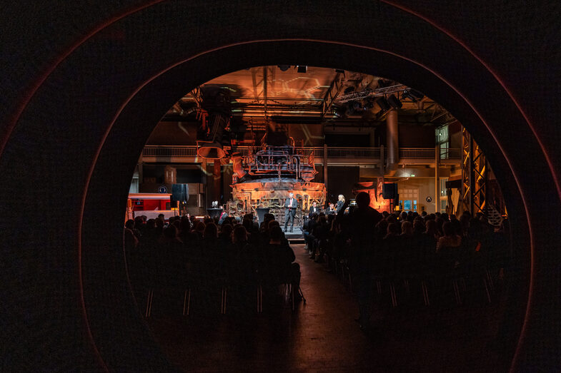 Foto einer illuminierten Halle. In dieser steht eine männliche Person auf einer Bühne und spricht zum Publikum, welches vor der Bühne sitzt.