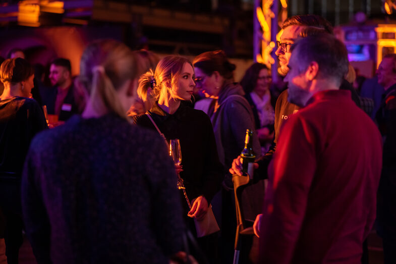 Mehrere männliche und weibliche Personen stehen in einer illuminierten Halle und unterhalten sich.