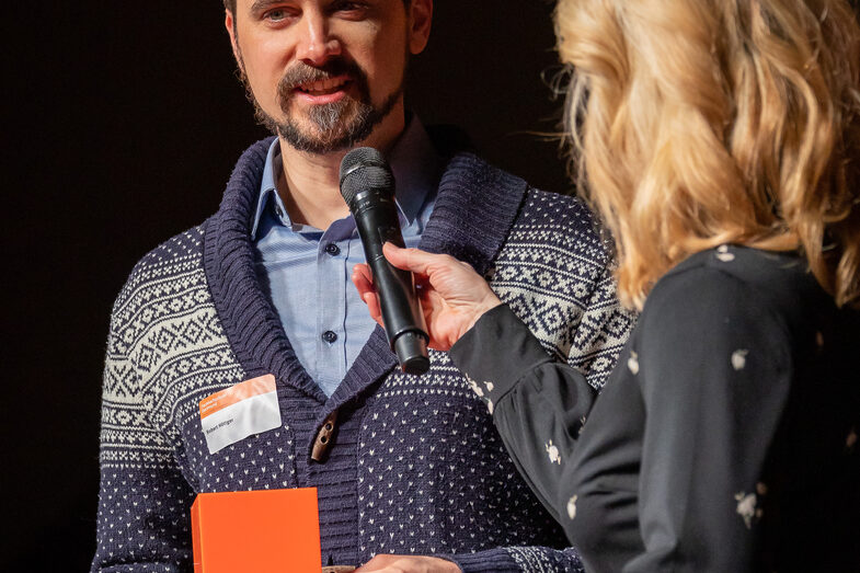 Foto von einer männlichen und einer weiblichen Person. Die männliche Person spricht in ein Mikrofon und hält dabei eine Mappe und einen Würfel in der Hand.