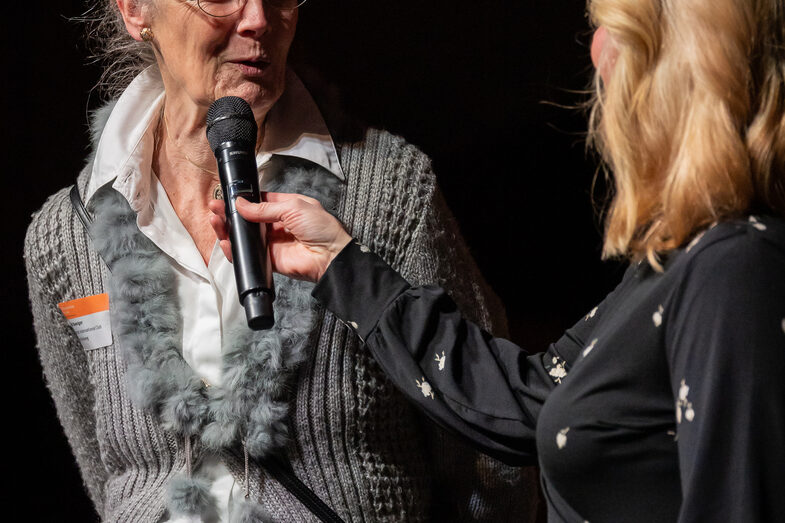 Foto von zwei weiblichen Personen. Eine Person spricht in ein Mikrofon.