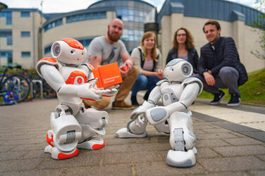 Photo of two NAO robots standing outside Building 42 and a group of students crouching behind them. Robot Eva holds an FH cube in her hand.