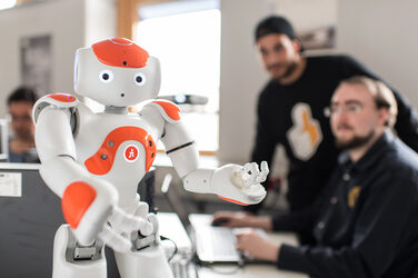 Foto von NAO-Roboter Eva, der vorne auf dem Tisch steht, Arme gehoben. Im Hintergrund zwei Männer, der eine am Computer, beide schauen zum Roboter. __ <br>NAO robot Eva stands in front of the table, arms raised. In the background someone is sitting at a laptop and talking to the person standing next to it. Both look at the robot.