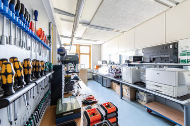 Room shot of the Hardeware workshop. You can see various tools on the workbench on the left and various printers on the right.